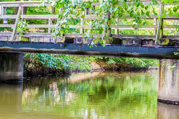 Small wooden footbridge over a stream, foliage with green leaves on railing, reflection on water surface, steel beams on two concrete pillars, sunny and calm spring day in the Netherlands 