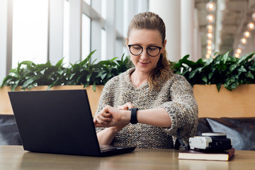 Wall Mural - Portrait of young businesswoman sitting in cafe,working on laptop,looks at wrist watch. Online marketing, education.