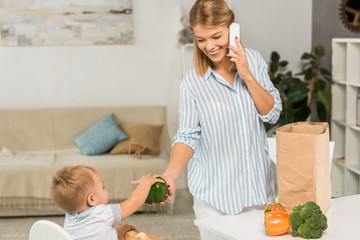 Wall Mural - happy mother holding groceries while talking on smartphone with toddler in baby chair on background