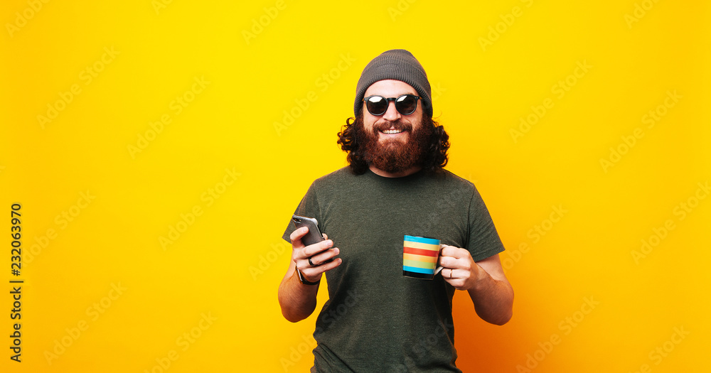 Happy bearded hipster man using phone and holding cup of coffee - obrazy, fototapety, plakaty 