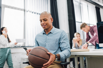 Wall Mural - smiling young african american businessman holding basketball ball in open space office