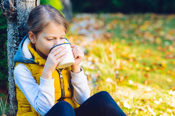 Wall Mural - child girl drinking chocolate from a cup dressed in a warm yellow vest in autumn scenery