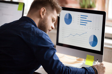 Back view of millennial male employee working on computer in office writing down stats results, focused worker make notes preparing report on pc, serious man busy making research analyzing graphs