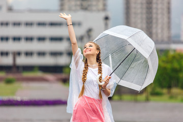 Young pretty girl with two braids in yellow boots and with transparent umbrella stands near fountain.