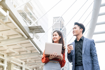 Beautiful businesswoman  holding laptop in her hands and colleague businessman holding coffee cup talking about work at outdoor street with building background.