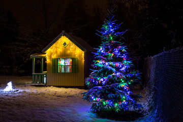 Wooden painted yellow private children`s play house in home garden, decorated with Christmas LED string lights outdoors in cold winder night. Decorated Christmas fir tree.