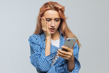Shocked Surprised Young Cute Woman With Strawberry Blonde Hair Looking At Her Phone, Receives Sms, News, Message, Isolated On Grey Background. Human Emotions, Reaction Concept