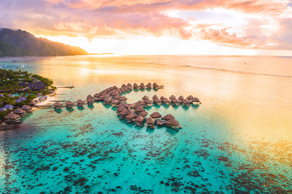 Luxury travel vacation aerial of overwater bungalows resort in coral reef lagoon ocean by beach. View from above at sunset of paradise getaway Moorea, French Polynesia, Tahiti, South Pacific Ocean. - obrazy, fototapety, plakaty 