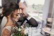 © sonyachny - Gorgeous bride and stylish groom gently hugging at window. Sensual wedding couple embracing. Romantic moments of newlyweds. Creative wedding photo through glass. Copy space