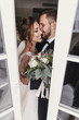 © sonyachny - Gorgeous bride and stylish groom gently kissing at window. Happy wedding couple embracing. Romantic moments of newlyweds. Creative wedding photo through glass. Copy space