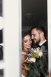 © sonyachny - Gorgeous bride and stylish groom gently hugging at window. Happy wedding couple embracing and smiling. Romantic moments of newlyweds. Creative wedding photo. Copy space