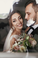 Wall Mural - Gorgeous bride and stylish groom gently hugging at window. Sensual happy wedding couple embracing and smiling in light. Romantic moments of newlyweds. Creative wedding photo