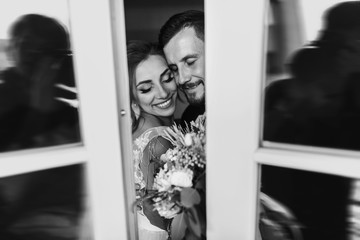 Wall Mural - Gorgeous bride and stylish groom gently hugging at window. Sensual wedding couple embracing. Romantic moments of newlyweds. Creative wedding photo through glass. Copy space