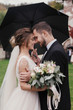 © sonyachny - Gorgeous bride and stylish groom gently hugging under umbrella in rainy outdoors. Sensual wedding couple embracing. Romantic moments of newlyweds. Modern wedding photo