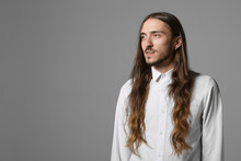 Beauty, Fashion, Style And People Concept. Isolated Shot Of Extraordinary Fashionable Young Male With Long Loose Hair, Beard And Earring Posing At Gray Wall, Wearing Stylish White Shirt, Smiling