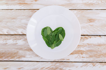 Wall Mural - Green leaves heart shape on white plate on rustic wooden background.