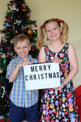 Wall Mural - Young children holding a Merry Christmas light up sign in front of the Christmas tree