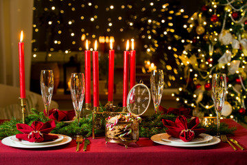 Beautiful served table with candles, Red tablecloth and napkins, white china, gold cutlery, crystal champagne glasses. Living room decorated with lights and Christmas tree. Holiday setting