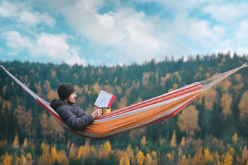 A man sits in a hammock and reads a book in a picturesque place. Mug in his right hand.