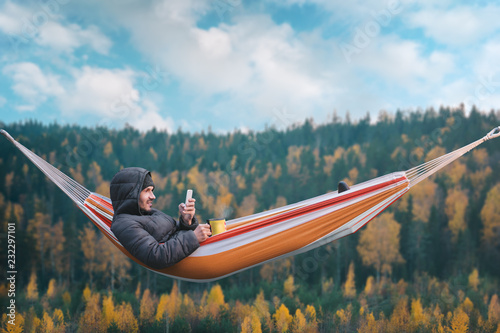 Photographie A smiling man sits in a hammock and uses a smartphone in a picturesque location