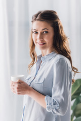 Wall Mural - happy adult woman holding cup of warming beverage and looking at camera