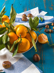 Orange ripe mandarin fruits with green leaves and chestnuts over rustic blue wooden background.