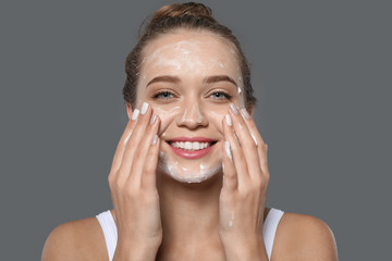 Young woman washing face with soap on grey background