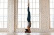 © fizkes - Young sporty woman practicing yoga, doing headstand exercise, salamba sirsasana pose, working out, wearing sportswear, pants and top, indoor full length, white yoga studio
