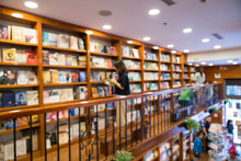 Blurred Abstract Background Of Bookshelves In Book Store, With A Girl Finding Book In The Store.
