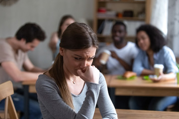 Diverse multi-ethnic friends sitting together in cafe talking having fun, focus on frustrated shy girl sitting separately by others teenagers feels unhappy because peers not accept her she is outcast