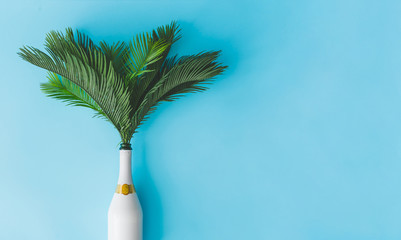 Wall Mural - Champagne bottle with tropical green palm leaves on pastel blue background. Summer holiday concept with creative copy space. Summer tropical celebration party flat lay.