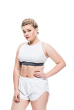 Fototapeta Dziecięca - young overweight woman in sportswear standing with hand on waist and looking at camera isolated on white