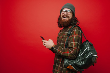 Cheerful smiling hipster man with beard using phone and wearing backpack over red background