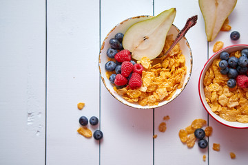 Golden cornflakes with fresh fruits of raspberries, blueberries and pear in two ceramic bowls. Prepared for healthy breakfast. Placed on white wooden table.