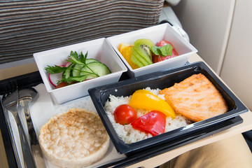 Food served in a passenger aircraft. In flight meal on the tray. Salad, butter and bread, fruits. Hot dish in the lunch box: salmon fish, rice and vegetables.