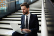 © pressmaster - Young mobile businessman sitting on stairs and looking at data on laptop display while networking
