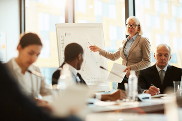 Wall Mural - Mature blond female coach pointing at graph on whiteboard and explaining it to audience at seminar