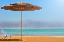 A Straw Umbrella And A White Chair Stand On A Sandy Beach Near The Water. Rest On The Dead Sea In Israel Overlooking The Mountains Of Jordan