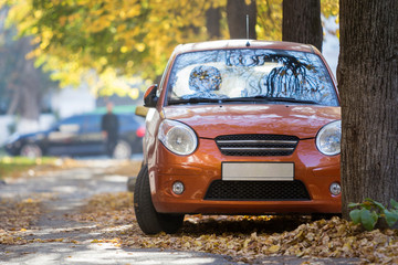 Wall Mural - Front view of small orange mini car parked in quiet yard on sunny autumn day on blurred buildings and big old trees golden foliage bokeh background. Transportation, parking problems concept.