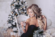 The blonde in a black dress with a glass sitting at the New Year's table looks to the side. Christmas tree in the background.