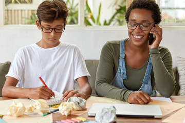 Wall Mural - Serious schoolboy in white t shirt, writes down records in notepad, busy with studying, cheerful African American lady has conversation via cell phone, controls work of her trainee, smiles broadly