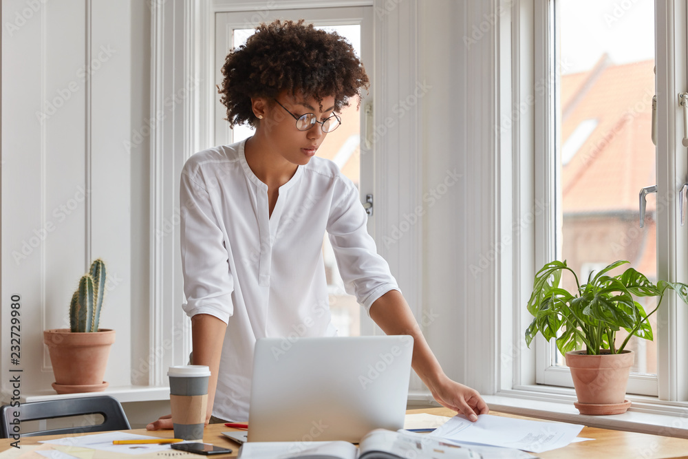 Horizontal shot of serious black young woman stands at desktop, analyzes income chart or data of finances, works with laptop computer, involved in business, wears white shirt and round spectacles.