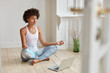 © wayhome.studio  - Calm lady with Afro hairstyle, dressed in sportswear, meditates on floor in empty room, listens spiritual practices lessons on laptop computer, poses in lotus pose, tries to relax. Yoga concept
