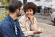 © WHstudio Leushin N - Mixed race woman and man look happily at each other, have rest in urban setting, eat fast food, have talk about something. Cheerful dark skinned girl holds delicious sandwich speaks with stranger
