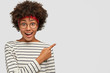 © wayhome.studio  - Studio shot of positive dark skinned girl wears round transparent glasses, striped white sweater, indicates with index finger at upper right corner, shows free space for advert. Promotion concept