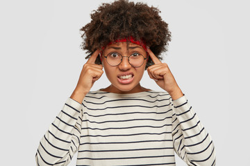 Wall Mural - Picture of displeased stressed dark skinned female teenager keeps both index fingers on temples, clenches teeth and frowns face with pain, dressed in striped sweater, models against white background