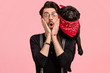 Emotive unshaven young man keeps both hands on cheeks, has his lovely black dog on shoulders, cant believe in shocking news, isolated over pink background. People, animals and reaction concept