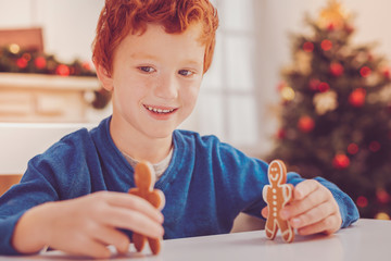 Wall Mural - Having fun. Pleasant upbeat red-haired boy sitting at the table, holding two gingerbread men and playing with them happily