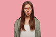 Indoor shot of indignant woman frowns face in displeasure, wears spectacles, shirt, models against pink background, doesnt like something dressed in casual shirt. Facial expressions concept.