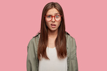 Indoor Shot Of Indignant Woman Frowns Face In Displeasure, Wears Spectacles, Shirt, Models Against Pink Background, Doesnt Like Something Dressed In Casual Shirt. Facial Expressions Concept.